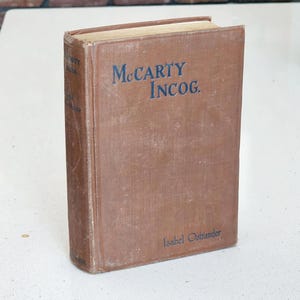 May include: A hardcover book titled McCarty Incog. by Isabel Ostrander with a brown cloth cover and dark blue lettering. The front cover displays the title in a serif font, and the author's name is printed in the lower right corner in blue.