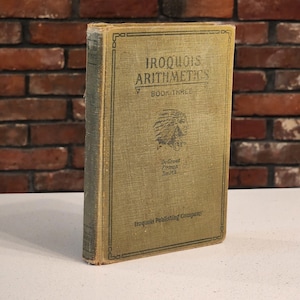 Puede incluir: Un libro antiguo titulado "Iroquois Arithmetics Book Three" con una cubierta verde descolorida. La portada presenta el título en negrita, una ilustración nativa americana y el nombre del autor. El libro está ligeramente desgastado.