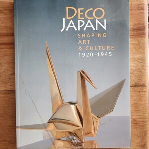 May include: A book titled "DECO JAPAN" with the subtitle "SHAPING ART & CULTURE 1920-1945." The cover features a gold origami crane sculpture against a gray background. The book is on a wooden surface.