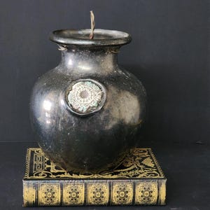 Puede incluir: Un jarrón oscuro de estilo antiguo con un sello decorativo. El jarrón es negro con una superficie texturizada y una abertura estrecha. Se encuentra sobre una caja decorativa con detalles dorados y negros. Una fina mecha se extiende desde la abertura del jarrón.