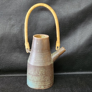 Può includere: Una brocca artigianale in ceramica con corpo marrone e base azzurra e verde. È dotata di un beccuccio e di un manico intrecciato in materiale marrone chiaro. La brocca viene probabilmente utilizzata per servire bevande.
