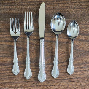 May include: A five-piece stainless steel flatware set consisting of a salad fork, a dinner fork, a dinner knife, a soup spoon, and a teaspoon. Each piece features a polished silver finish and an ornate, embossed scroll pattern on the handle.