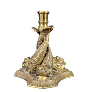 May include: A brass candlestick holder featuring a design of three fish. The fish are intertwined, supporting the candle cup. The base is a decorative, multi-sided shape. The candlestick is made of brass and has a gold color.