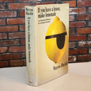 May include: A vintage hardcover book titled "If you have a lemon, make lemonade" by Warren Hinckle. The cover features a yellow lemon with a black eye mask. The book's spine also displays the title. The background is a brick wall.