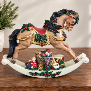 Könnte beinhalten: Eine beige Schaukelpferd-Figur mit schwarzer Mähne und Schweif, verziert mit grünen Stechpalmenblättern und roten Beeren. Der Sockel zeigt einen Geschenkkorb, eine kleine Puppe im roten Kleid, eine Trommel und eine Gitarre auf einem cremefarbenen Bogen.