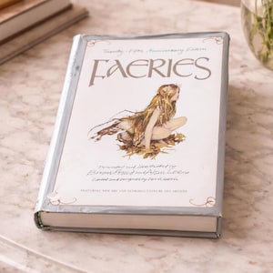 May include: A hardback book titled "FAERIES" with a light grey cover and silver trim. The cover features an illustration of a seated woman with long hair and a flowing garment. The text includes "Twenty-Fifth Anniversary Edition".