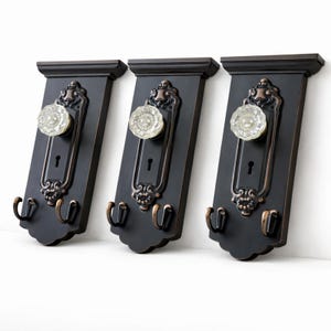 May include: Three black wall-mounted coat racks with bronze-coloured ornate detailing. Each rack has a clear glass doorknob and two curved hooks. The racks have a vintage, antique aesthetic.