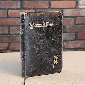 Puede incluir: Un libro pequeño de tapa dura negra con el título Following of Christ en letras doradas en la portada. La cubierta presenta un emblema de cruz dorada y muestra signos de desgaste. El lomo también muestra el título en letras doradas.