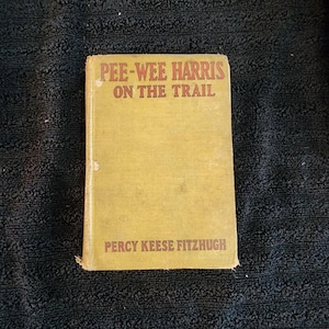 Puede incluir: Un libro antiguo titulado "Pee-Wee Harris on the Trail" de Percy Keese Fitzhugh. El libro tiene una cubierta amarilla con letras rojas. El libro está ligeramente desgastado, lo que sugiere que es una edición antigua.
