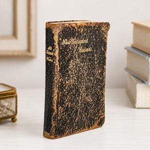 May include: A vintage, weathered book titled New Testament and Psalms with a textured black and gold mottled cover. The spine and front cover feature gold embossed lettering. The edges of the cover show signs of wear with exposed brown material.