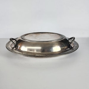 May include: A silver-colored serving dish with a lid. The dish is oval-shaped with a raised rim and two handles. The lid has a decorative rope-like border. The dish is likely made of metal and is designed for serving food.