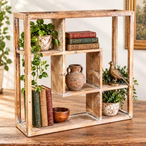 May include: A square wooden shelving unit with a distressed finish, featuring multiple compartments. It holds a potted green plant, stacks of hardcover books, a brown ceramic vase, a small wooden bowl, and a metallic bird figurine.