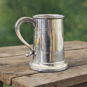 May include: A polished silver tankard with a curved handle. The tankard has a wide base and a slightly flared top. It is displayed on a rustic wooden surface.