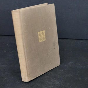 Peut inclure: Un livre à couverture rigide avec une couverture et un dos de couleur beige. Un carré décoratif doré est centré sur la couverture. Le livre est légèrement incliné, révélant le dos et le bord des pages.