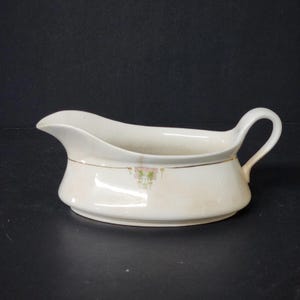 May include: Cream-colored gravy boat with a curved spout and a handle. The boat features a floral design in pink and green, with a gold trim. The gravy boat is made of ceramic and is likely vintage.