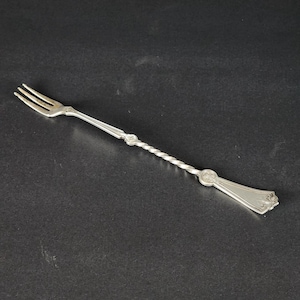 May include: A silver serving fork with a three-pronged head and a twisted handle. The handle has decorative elements near the head and the base. The fork is made of a shiny, reflective metal.
