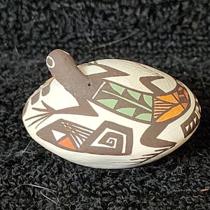 May include: Hand-painted ceramic turtle figurine. The turtle has a brown head and legs, with a cream-coloured shell decorated with geometric patterns in brown, orange, and green. The figurine is resting on a dark, textured surface.