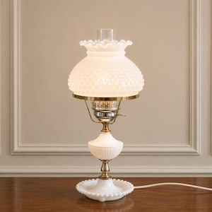 May include: A table lamp featuring a white milk glass hobnail shade with a ruffled top edge, a clear glass chimney, and a brass-toned metal burner assembly. The base is constructed from white milk glass with a fluted design and a circular foot.