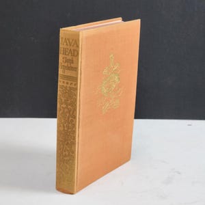May include: A hardcover book titled Java Head by Joseph Hergesheimer with a light orange cloth cover. The spine features gold embossed text and floral scrollwork, while the front cover displays a central gold floral emblem and decorative details.