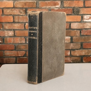 May include: A vintage hardcover book titled Smyth's Elements of Algebra with a black spine and a textured gray cover. The title is printed in gold lettering on the spine, which shows signs of wear along the edges and corners of the binding.