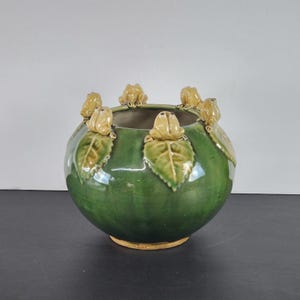 Puede incluir: Un jarrón redondo de cerámica verde con un acabado brillante. El jarrón está adornado con figuras de ranas de color beige claro posadas sobre acentos de hojas verdes alrededor del borde. La base del jarrón es de color marrón claro.