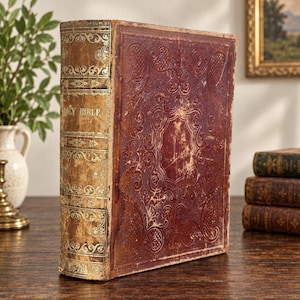 May include: An antique Holy Bible with a deep red, embossed leather cover featuring intricate scrollwork patterns. The spine is light brown with gold-toned decorative detailing and the text "HOLY BIBLE" clearly printed in gold lettering.