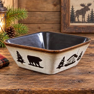 May include: A square ceramic bowl with a glossy dark interior and a cream-coloured exterior. The outer sides feature a dark brown decorative pattern depicting silhouettes of pine trees, a walking bear, and a small cabin in a rustic style.