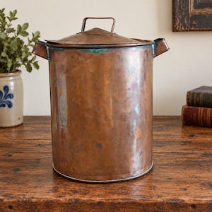Könnte beinhalten: Ein zylindrischer Kupferbehälter mit passendem Deckel und zwei seitlichen Griffen. Die Metalloberfläche zeigt einen warmen, polierten Kupferton mit sichtbaren Bereichen natürlicher Oxidation und blau-grüner Patina am oberen Rand.