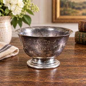 May include: A tarnished silver-toned metal bowl with a pedestal base. The exterior surface features the engraved text "EASTCO" above the year "1972". The bowl has a wide, flared rim and a weathered, dark patina finish across its entire exterior.