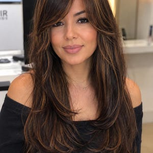 May include: A long, layered wig featuring dark brown hair with warm caramel highlights. The hair is styled with side-swept bangs and soft, face-framing layers that cascade down past the shoulders, creating a voluminous and textured appearance.