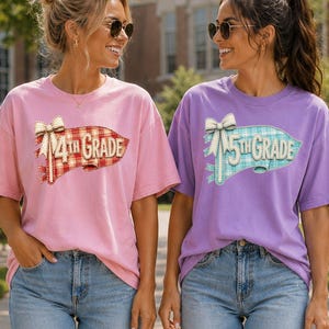 May include: A light pink t-shirt with a red plaid pennant graphic reading 4th GRADE and a light purple t-shirt with a blue plaid pennant graphic reading 5th GRADE. Both shirts feature a decorative bow above the text and are paired with blue jeans.
