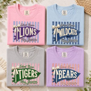 May include: Four crewneck t-shirts in pink, light blue, grey, and lavender. Each features a graphic of a pennant with a bow, the text School Name, a team name (LIONS, WILDCATS, TIGERS, BEARS), and the text Mrs. Jessica printed on the front.