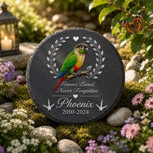 May include: A round black slate memorial stone featuring a colorful green, yellow, and red parrot perched on a branch. The stone includes a silver laurel wreath, a heart, the text Forever Loved, Never Forgotten, the name Phoenix, and 2010-2024.