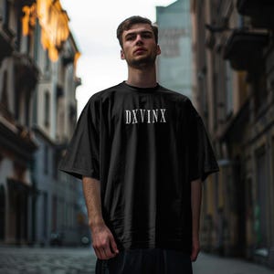 Könnte beinhalten: Ein schwarzes Kurzarm-T-Shirt mit dem in Weiß auf die Brust gedruckten Text DXVINX. Das Kleidungsstück hat einen Rundhalsausschnitt und eine lockere, entspannte Passform mit weiten Ärmeln, gefertigt aus einem einfarbigen schwarzen Stoff mit glatter Textur.