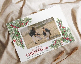 May include: A Christmas card with a watercolor border of holly and greenery. The card features a photo of a family of three playing in a field. The text on the card reads "Merry Christmas" and "The Thompson Family, 2022".