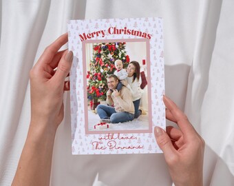 May include: A Christmas card with a photo of a family of three standing in front of a decorated Christmas tree. The card has a pink and white background with a pattern of small Christmas trees. The text "Merry Christmas" is written in red at the top of the card, and the text "with love, The Dinnams" is written in red at the bottom of the card.