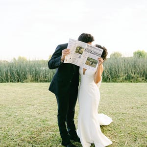 Może przedstawiać: Para stoi na polu, trzymając gazetę przed twarzami. Gazeta nosi tytuł "The Wedding News" i zawiera zdjęcie pary ślubnej. Mężczyzna jest ubrany w czarny garnitur, a kobieta w białą suknię ślubną.