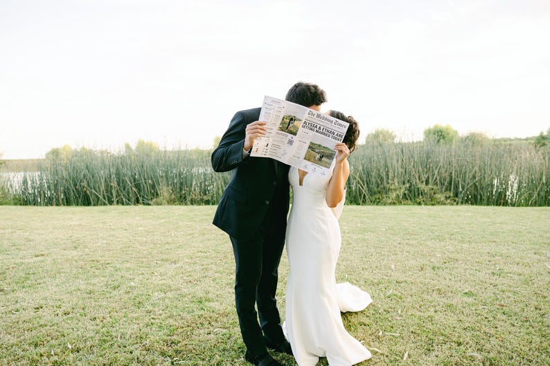 Może przedstawiać: Para stoi na polu, trzymając gazetę przed twarzami. Gazeta nosi tytuł "The Wedding News" i zawiera zdjęcie pary ślubnej. Mężczyzna jest ubrany w czarny garnitur, a kobieta w białą suknię ślubną.
