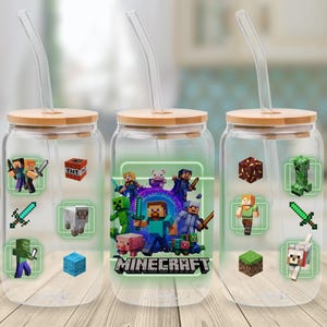 Könnte beinhalten: Drei transparente Glasbecher mit Bambusdeckeln und gebogenen Glasstrohhalmen. Jeder Becher zeigt Minecraft-Grafiken, darunter Charaktere, Blöcke und das Minecraft-Logo. Die Becher stehen auf einer Holzoberfläche.