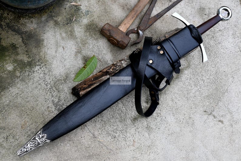 Op de afbeelding: Een zwarte leren schede met een zilverkleurig zwaard erin. De schede heeft een riem en een gesp. Het zwaard heeft een zilverkleurig handvat en een zwarte handgreep. Het zwaard ligt op een grijs betonnen oppervlak.