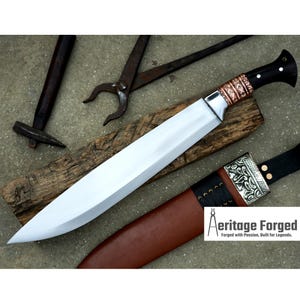 May include: A long, silver blade with a brown leather sheath and a copper and black handle. The blade is resting on a wooden surface. The text "Heritage Forged" is visible in the lower right corner of the image.  Forged with Passion, Built for Legends.