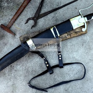 May include: A black and gold sword with a leather sheath and strap, resting on a wooden block. The sword has a curved blade and a decorative handle. A hammer and pliers are in the background.