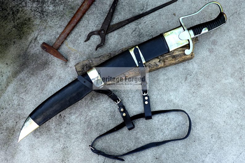 May include: A black and gold sword with a leather sheath and strap, resting on a wooden block. The sword has a curved blade and a decorative handle. A hammer and pliers are in the background.