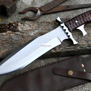 Puede incluir: Un cuchillo grande de hoja fija plateado con mango de madera marr&oacute;n. El cuchillo tiene una funda de cuero con una correa. El cuchillo est&aacute; sobre una superficie de madera con un objeto met&aacute;lico en el fondo. El texto "Heritage Forged" est&aacute; impreso en la hoja.