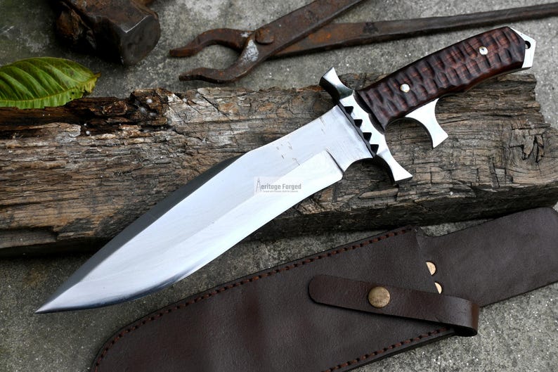 Puede incluir: Un cuchillo grande de hoja fija plateado con mango de madera marr&oacute;n. El cuchillo tiene una funda de cuero con una correa. El cuchillo est&aacute; sobre una superficie de madera con un objeto met&aacute;lico en el fondo. El texto "Heritage Forged" est&aacute; impreso en la hoja.