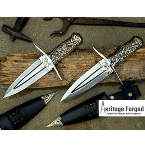May include: Two ornate daggers with silver blades and gold-toned handles with intricate Celtic knot designs. The daggers are displayed on a wooden surface with leather sheaths.