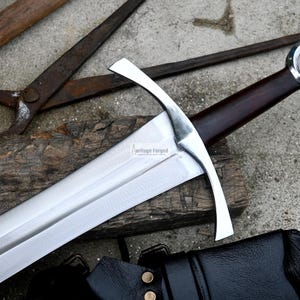 Op de afbeelding: Een zilveren middeleeuws zwaard met een bruine houten handvat en een ronde pommel. Het zwaard rust op een stuk hout met een zwarte leren schede op de achtergrond.