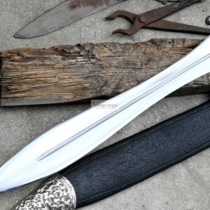 May include: A silver sword with a black leather scabbard and a wooden handle. The sword has a pointed tip and a double-edged blade.