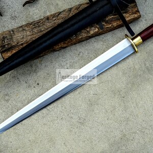 May include: A long, silver blade sword with a dark wood handle and gold accents. The sword is displayed with a black sheath and rests on a wooden plank. The text "Aeritage Forged" is visible on the blade.