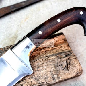 May include: A close-up of a large, silver knife with a dark wood handle. The handle has white dots and a gold ring. The knife is resting on a piece of wood.
